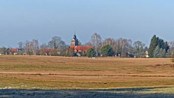 Blick auf die Basdorfer Kirche über eine sonnendurchflutete Wiesenlandschaft im Winter hinweg.